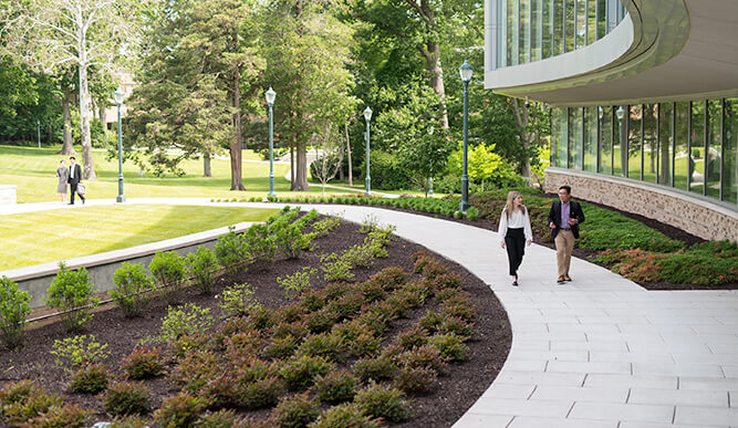 Two individuals stroll along a walkway adjacent to a the Dolan School of Business building, enjoying their surroundings in a serene atmosphere.  